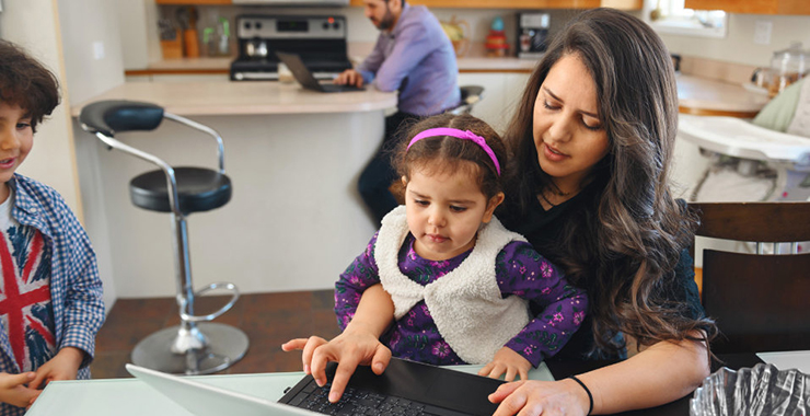 woman with a child on her lap, at her kitchen counter on a laptop