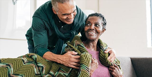 an older man wrapping a blanket over the shoulders of an older woman