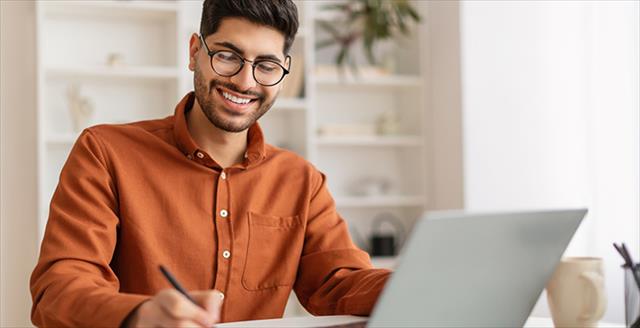 man writing on a piece of paper while sitting in front of a laptop