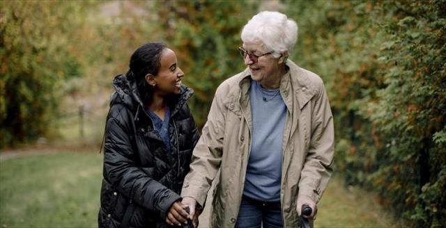 Caregiver smiling and assisting an older woman on a walk through the park.