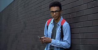 outcast African American male teen using cellphone outside school building