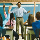 Black male teacher standing in front of a class of elementary school students raising their hands