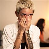 Young woman sits on bed, clasping her hands over her face in exasperation.