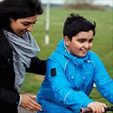 Young child with autism rides a bike while being supported by his mom, who has her hand on his upper back.