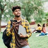 A young person holding a phone and notebook stands on a grassy campus, looking into the distance with other students seated in the background.