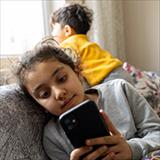 A young girl sitting on a couch, engrossed in her cell phone while her little brother stands behind her