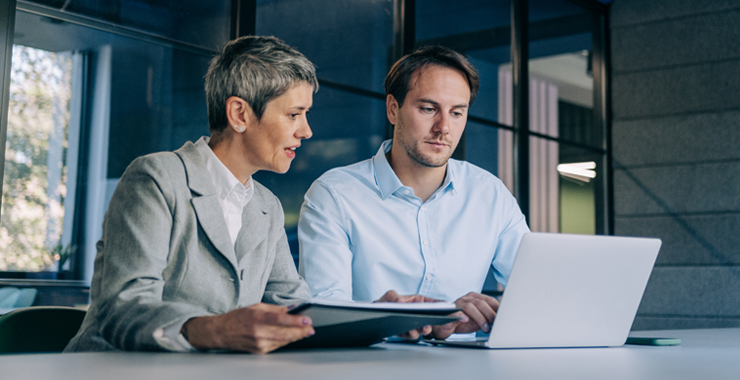 Two professional people speaking to each other while working on a laptop.