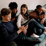 Four teenagers of various races and ethnicities relaxing on a couch while watching videos on their smart phones.