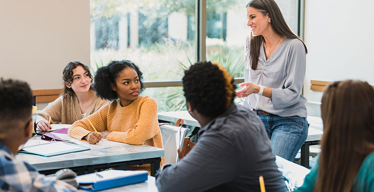 High school teacher talks with students at desk.