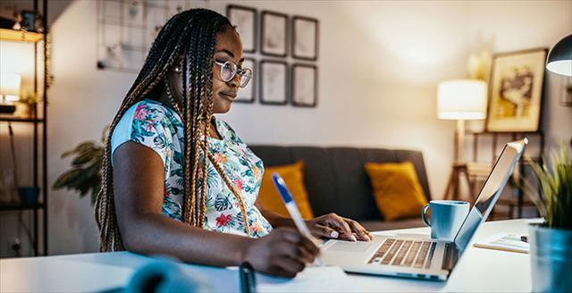 African American woman writes in notebook while looking at laptop screen.