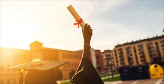 This image shows a person celebrating their graduation by holding up a diploma at sunset
