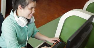 Young, blind student using a computer while sitting at a desk with headphones around her neck.