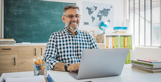 Older male teacher sits at desk in classroom using laptop.