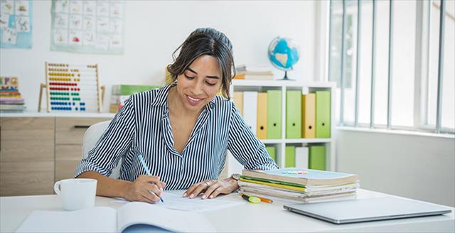Young female teacher sits at desk in classroom writing notes on a notepad.