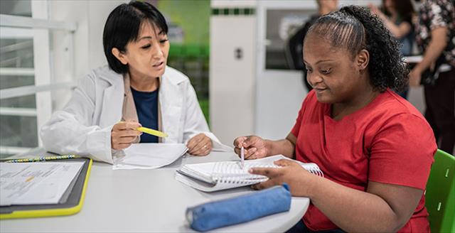 Older, Asian female teacher assists young, African American student who has a neurodevelopmental disability.