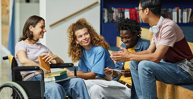 Racially diverse group of college-aged students, including one seated in a wheelchair, conversing in the library.