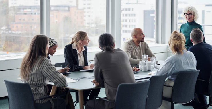 A group of coworkers sit around a conference table in a bright, modern meeting room, engaged in discussion with notebooks, cups, and city windows in the background.