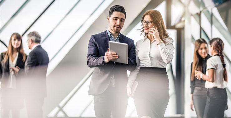 Professionals walk through a bright hallway while discussing work on a tablet and phone.