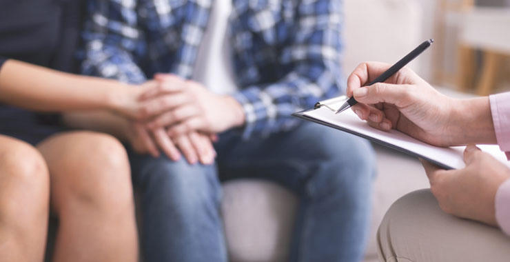 A therapist holding a clipboard takes notes while two people sit side by side during a counseling session.