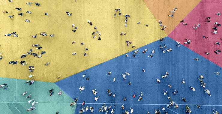 Overhead view of dozens of people walking across a large street painted in bold multicolor geometric blocks.