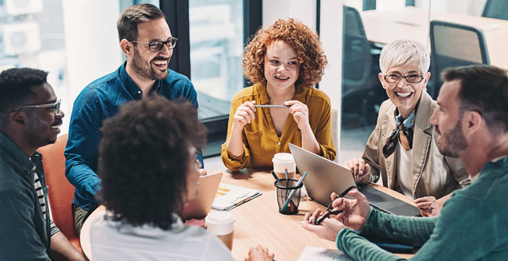 A group of coworkers sits around a conference table, engaging in discussion with laptops, notebooks, pens, and coffee cups in a bright office meeting space.