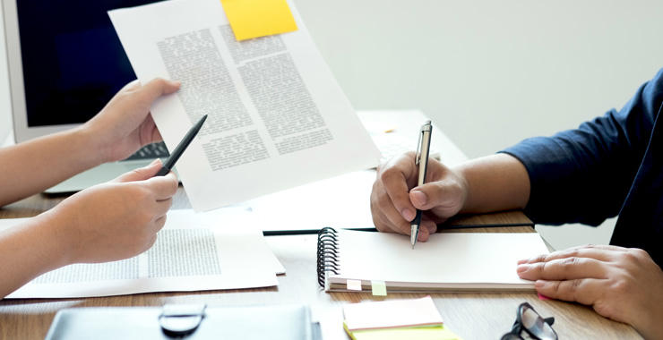 Two individuals’ hands review printed documents at a desk, with one holding a pen over a notebook while the other points to a page marked with a sticky note.