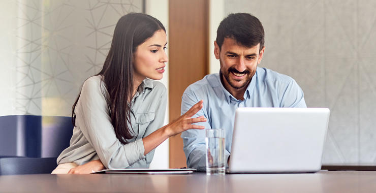 Two coworkers collaborate at a table, with one gesturing toward an open laptop while the other looks on, alongside a glass of water and a tablet.