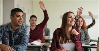 College students seated in classroom smiling, with hands raised.