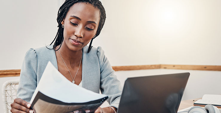 African American woman looks over report while sitting in front of her laptop.