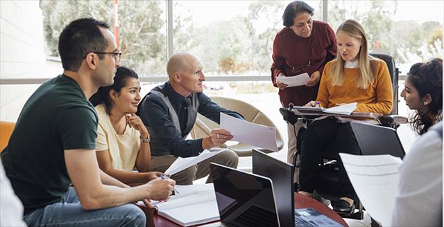 A multi-generational group of researchers gathered around a table in a light-filled workspace having an intense discussion we can assume is about research they are collectively passionate about