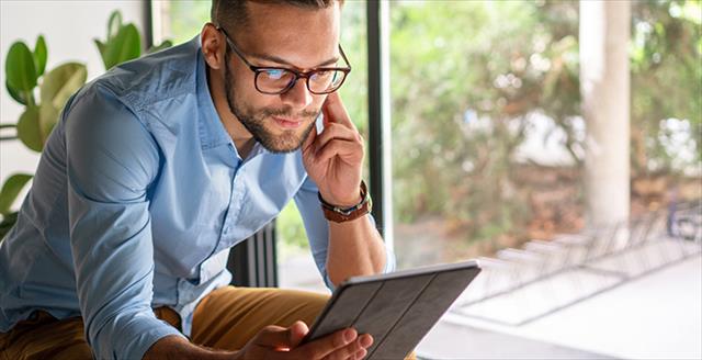 Man sits in a light-filled room pensively reading a tablet while the screen reflects in the glasses he wears to look smarter