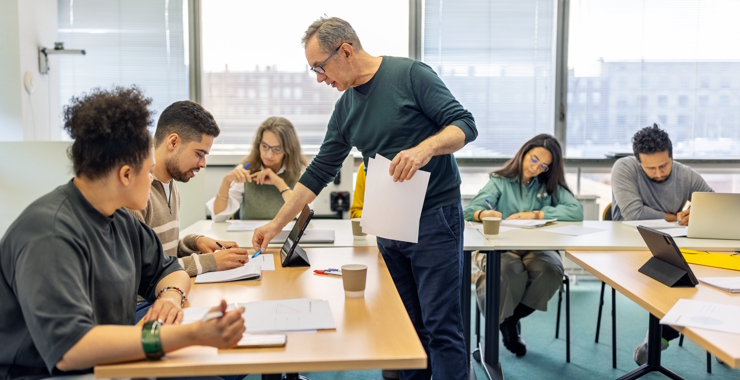 Senior professor talks with male student sitting at desk in adult education classroom