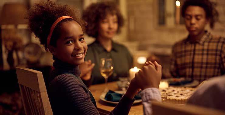Black girl smiling at camera as her family prays over a meal