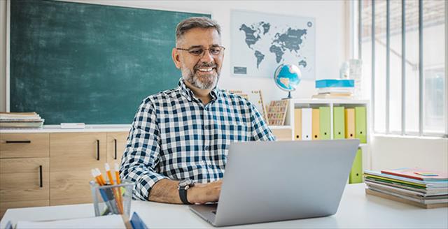 Older male teacher using laptop at desk in classroom.