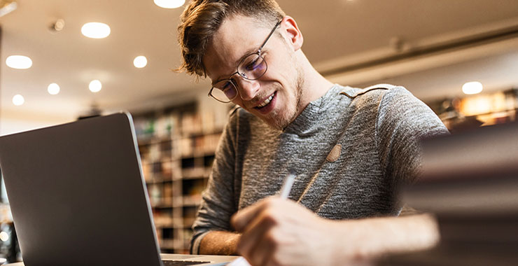 Male college student takes notes while working on laptop.