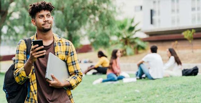 A young man holding a phone and notebook stands on a grassy campus, looking into the distance with other students seated in the background.