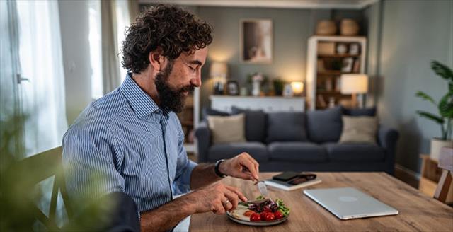 A man seated at a dining table eating a plate of salad, with a closed laptop nearby.