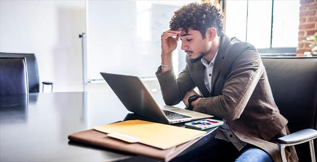 Man at a conference table working on a laptop, hand on forehead, with documents nearby.