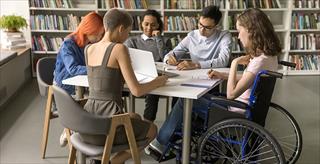 Group of college students, including one student in a wheelchair, sit together in the library.