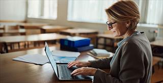 Teacher seated at desk using a laptop.