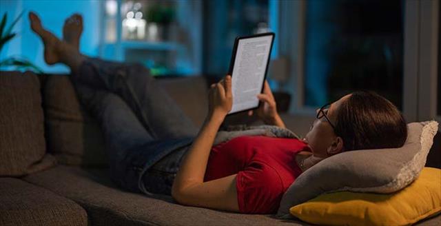 Woman relaxing on a couch at home late at night, feet up, reading digital content on her tablet