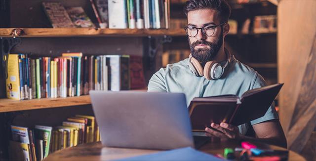 Researcher sitting in a warm library office setting reading out of a book and working on a laptop with focused determination