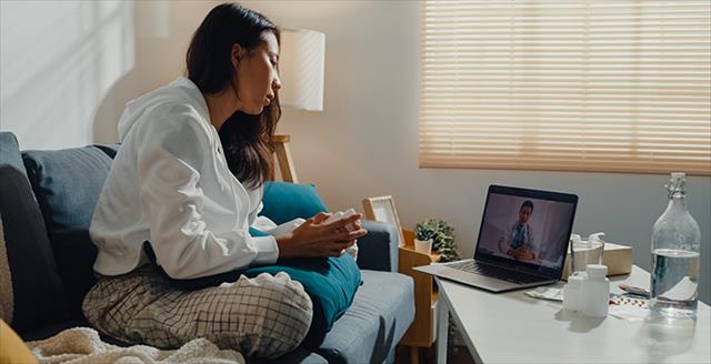 Sick young Asian woman using laptop talk to doctor sitting on sofa in living room at home