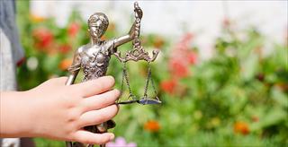 Kid's hand holding sculpture of themis, femida or justice goddess on green leaves natural bokeh background