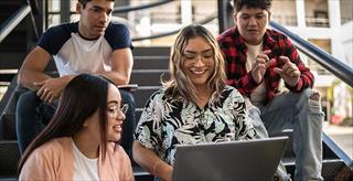 Two female students sit in front of two male students on the steps of their college campus; one female student holds a laptop to research for the group.
