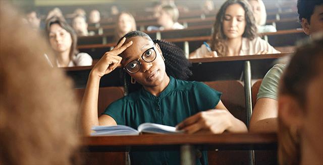 Young college student seated in lecture hall with overwhelmed expression on face.
