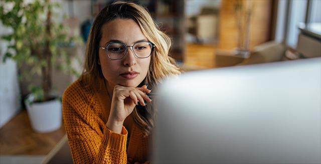 Studious young woman with glasses gazes with fascination and interest at what she is observing on her desktop screen while the surrounding ambience gives moody, cozy autumnal vibes