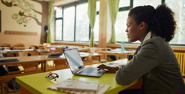Young teacher using laptop at desk in classroom.