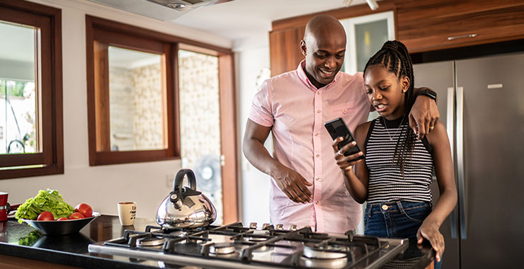 African American father and daughter standing in kitchen looking at daughter's phone screen.