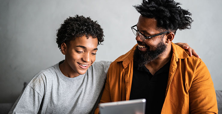 Older African American man laughs alongside young African American boy; both looking at tablet screen.